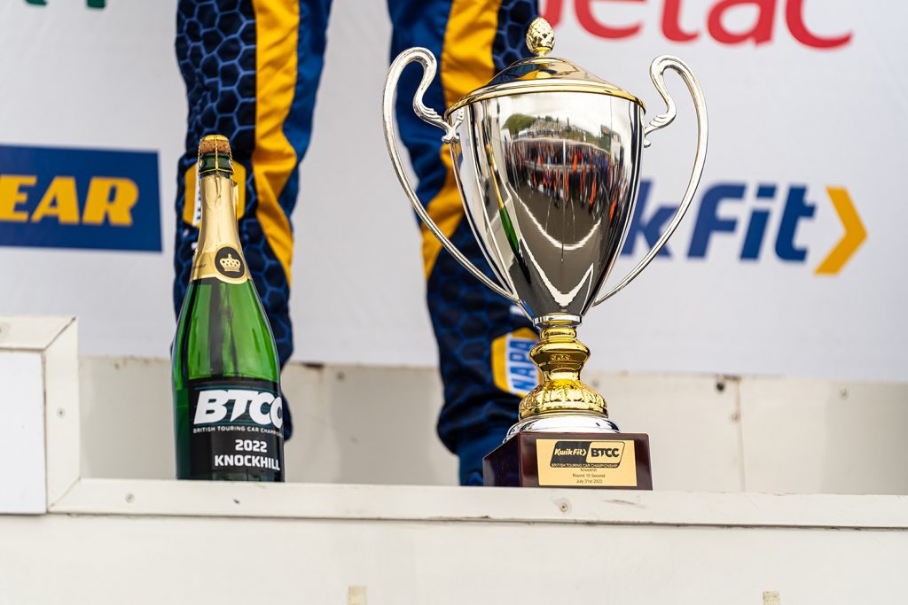 Ash Sutton stands proudly on the first place podium at BTCC Knockhill with the first place trophy and a bottle of champagne at his feet.