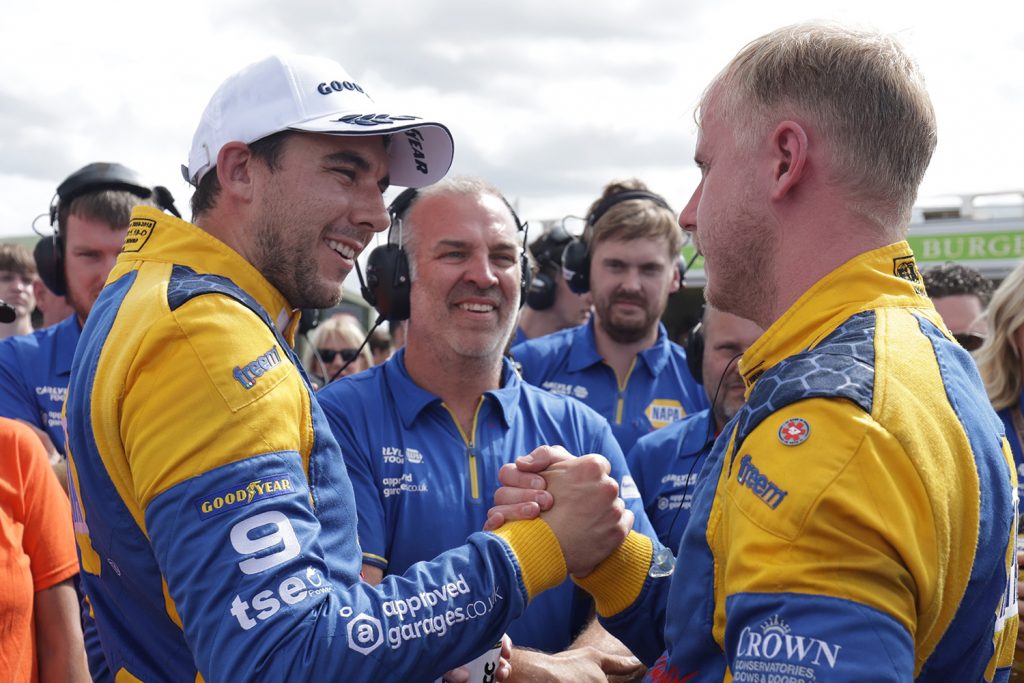 Dan Cammish and Ash Sutton shake hands with bright smiles in their NAPA overalls on a BTCC race day.