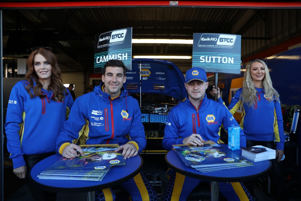 NAPA Racing UK's Dan Cammish (left) and Ash Sutton (right) sit at their BTCC autograph tables smiling at the camera with their grid girls standing on either side.